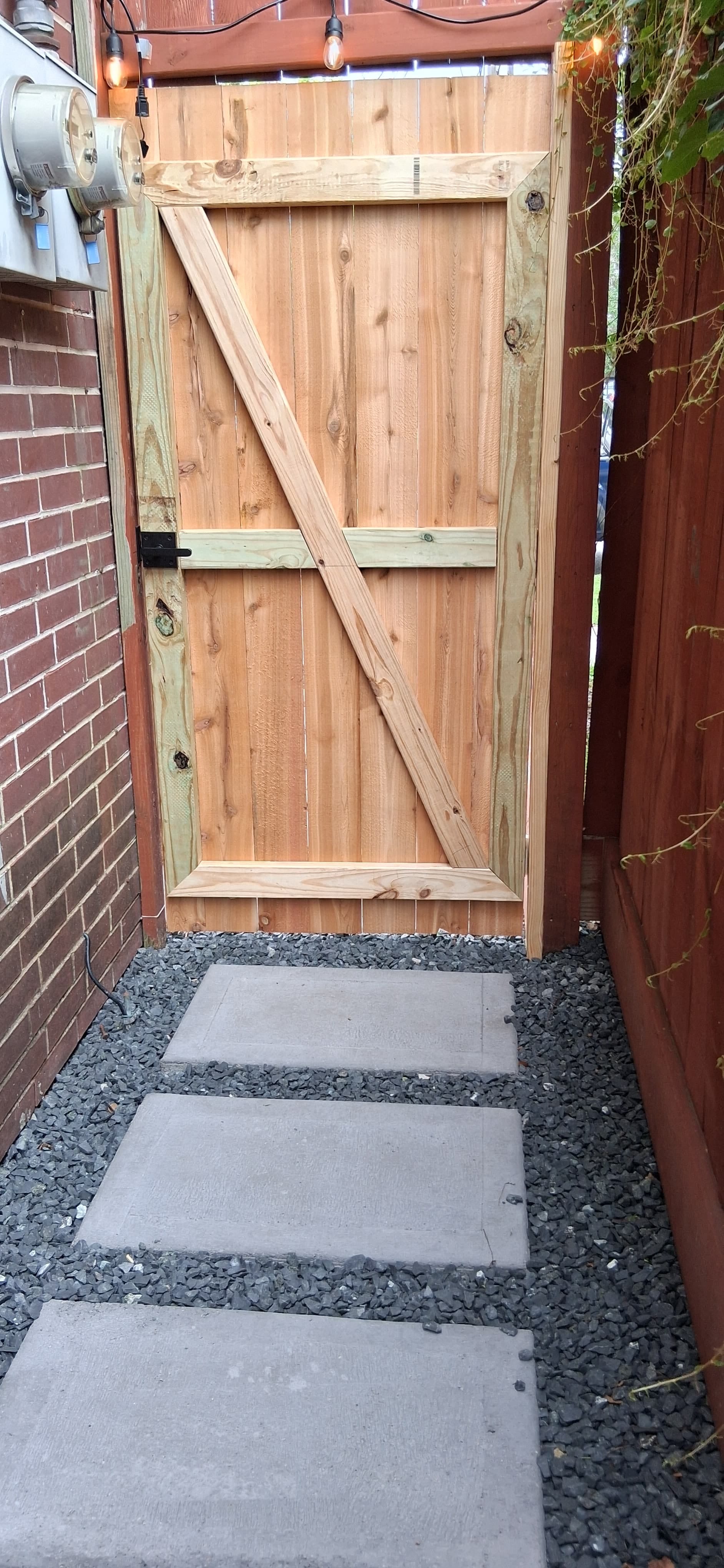 Completed wood gate with diagonal bracing and black hardware at the end of a concrete walkway.