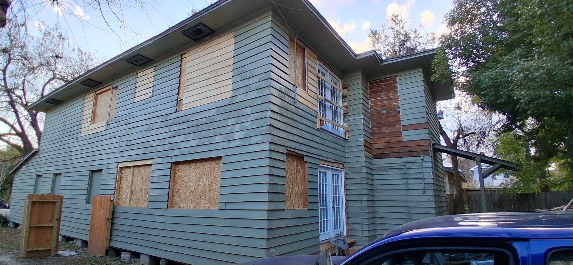 Side and rear of the same house with boarded window openings and exposed siding repair areas before window and trim installation.