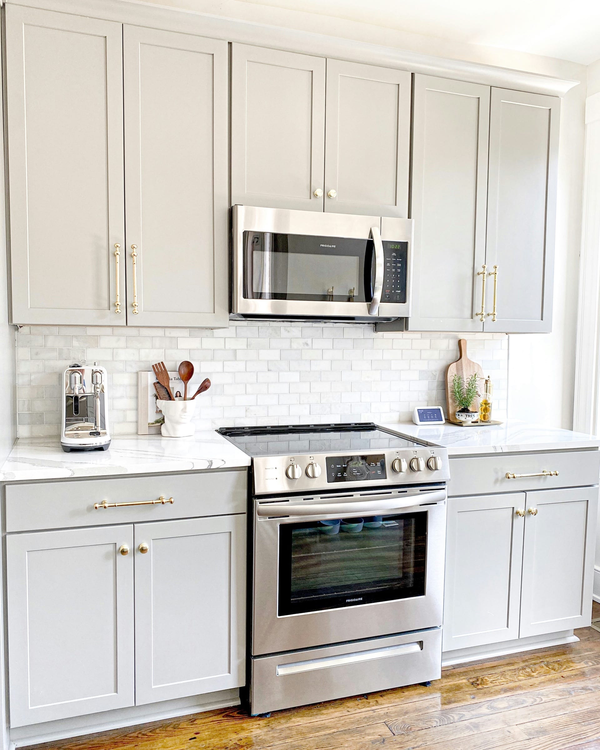 Light gray shaker cabinets with brass knobs and pulls, a white tile backsplash, and a stainless steel range.