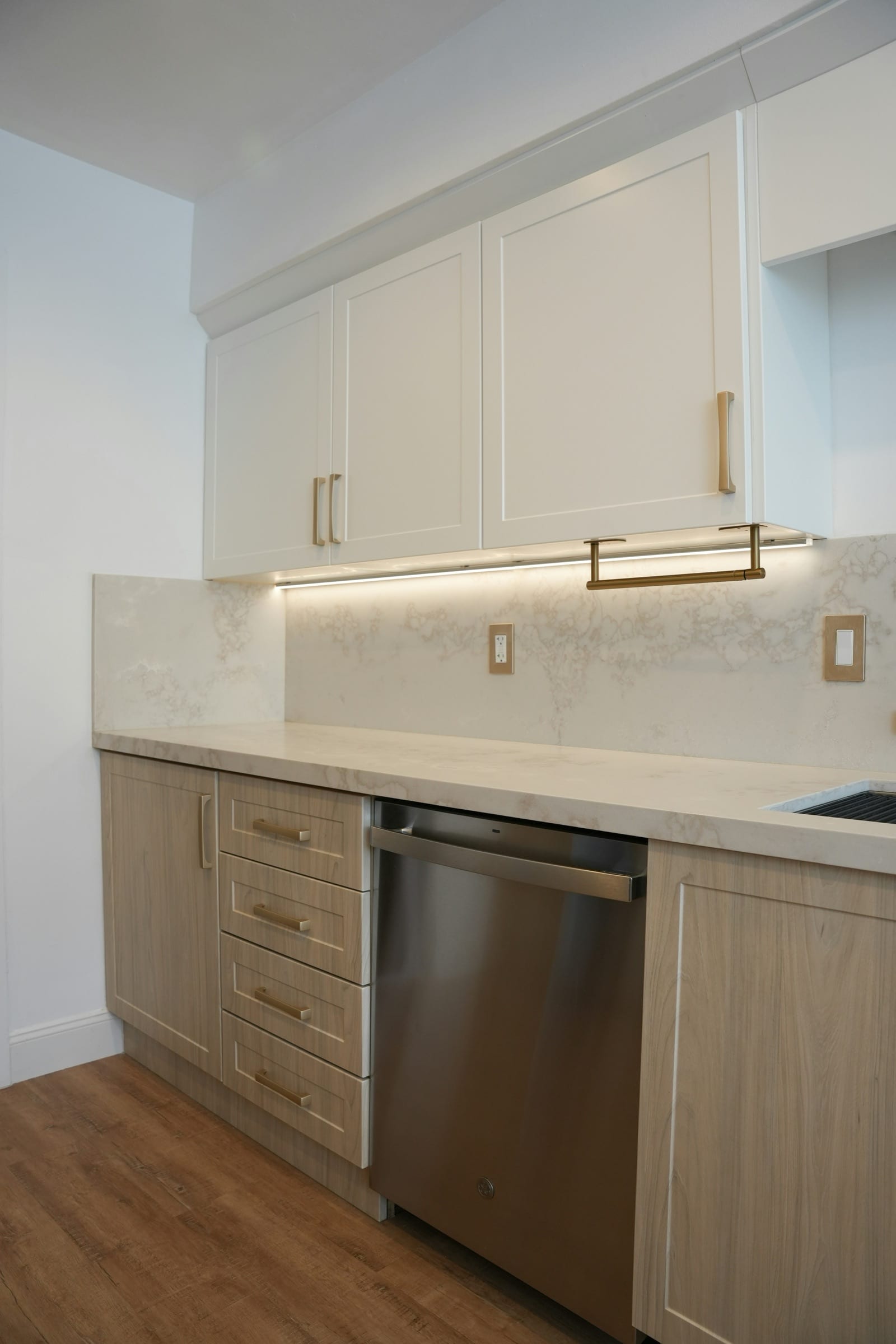 White upper cabinets and light wood-tone lower cabinets with brass pulls, a pale backsplash, and a stainless steel dishwasher.